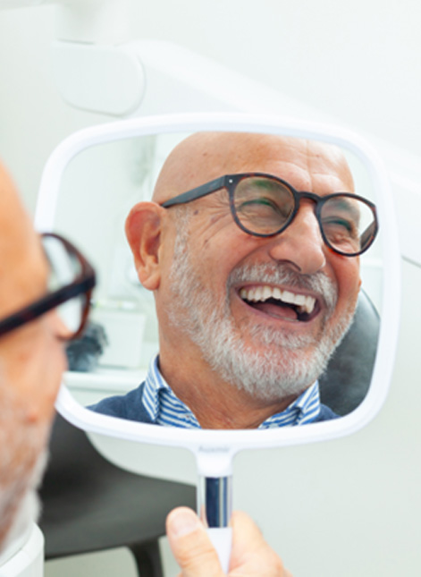 Senior man with glasses looking at handheld mirror in dental office